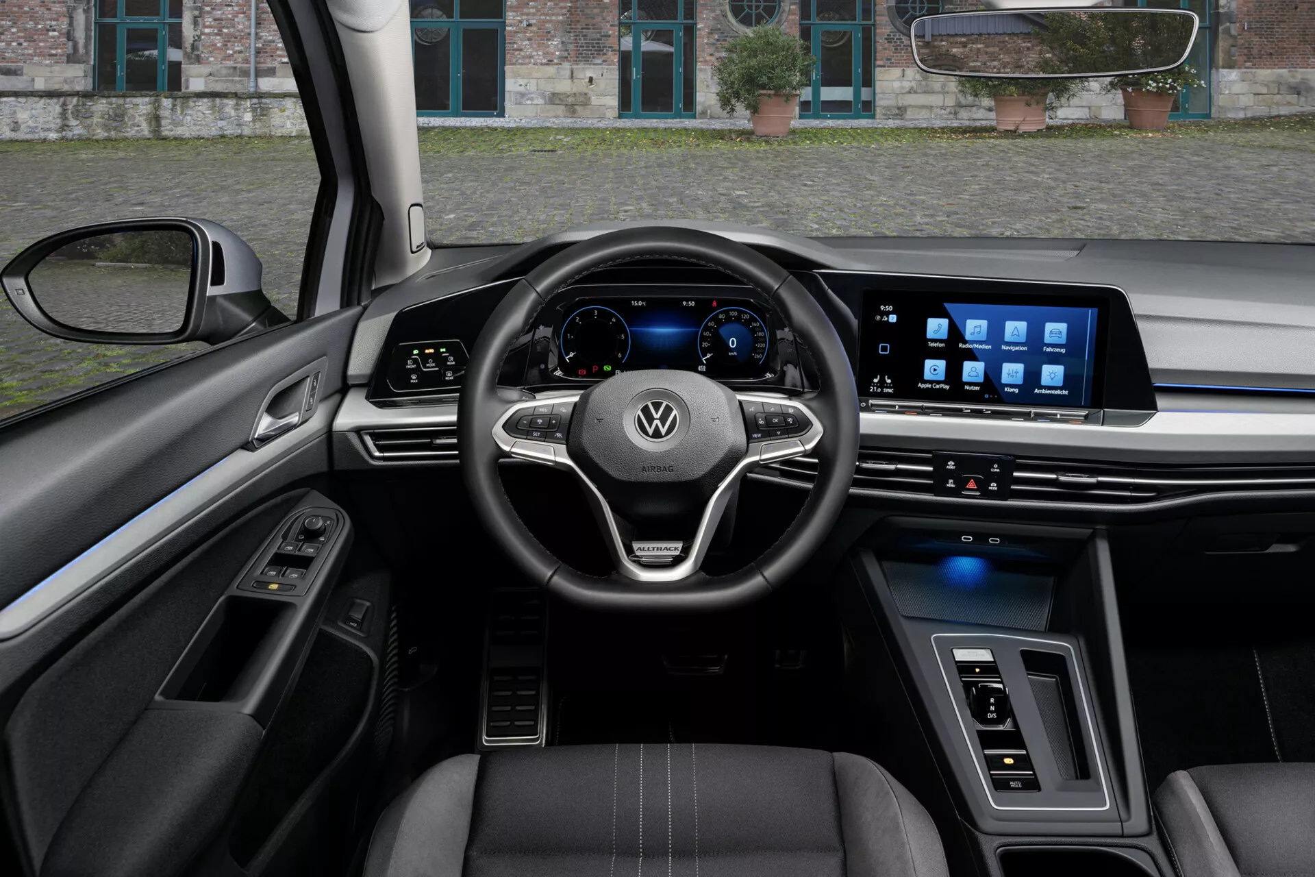 Vista interior de um VW Golf Alltrack 2017 sob a perspectiva do motorista, exibindo um volante de três raios com controles montados e logotipo Volkswagen, um cluster de instrumentos digital e um amplo sistema de infoentretenimento touchscreen no painel, destacado por recursos tecnológicos avançados, assentos revestidos em couro com costura contrastante, e uma mistura de materiais de luxo e iluminação ambiente.