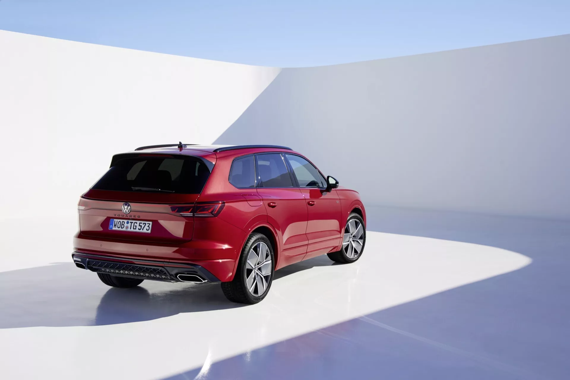 Modelo VW Touareg III facelift 2023 fotografado a partir de uma vista traseira de três quartos destacando o lado direito, em estúdio com fundo contínuo, exibindo seu acabamento vermelho brilhante, design aerodinâmico com linhas de carroçaria acentuadas, luzes traseiras LED, limpador de vidro traseiro, spoiler montado no teto e rodas de liga leve.