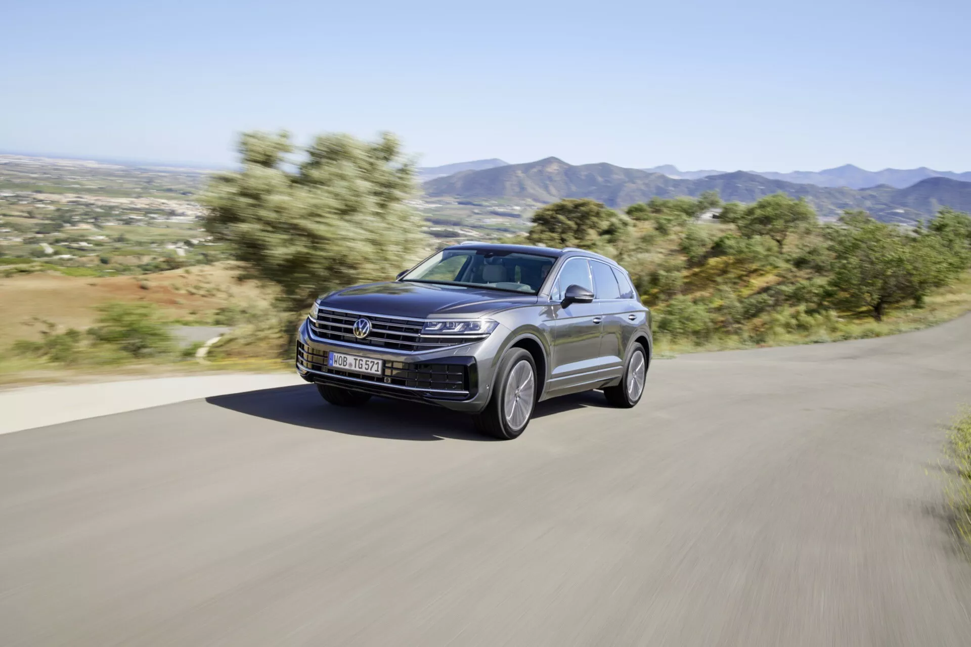 Modelo VW Touareg 2023 facelift dirigindo dinamicamente em uma estrada rural pavimentada, exibindo seu design moderno e características de desempenho, incluindo faróis LED, pintura metálica de luxo e rodas de liga leve elegantes, com um pano de fundo montanhoso